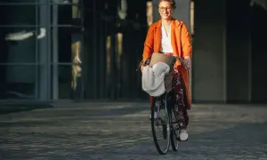 Uśmiechnięta kobieta w pomarańczowym płaszczu jedzie rowerem do pracy. W koszyku ma torbę i biały szal.