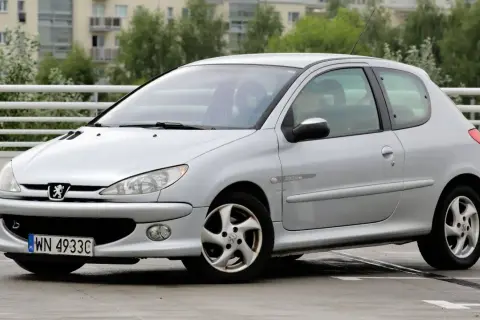 Srebrny Peugeot 206 z widocznymi przednimi żarówkami, zaparkowany na mokrym parkingu.