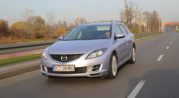 Fioletowa Mazda 6 GH jedzie po drodze. Jej żarówki świecą jasno, oświetlając trasę.