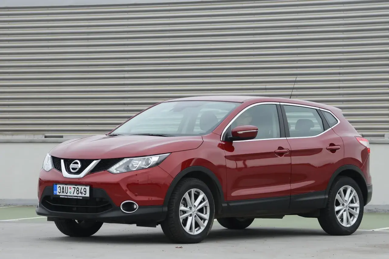 Czerwony Nissan Qashqai J11, z widocznym wykazem żarówek w przednich reflektorach, zaparkowany na tle falistej ściany.