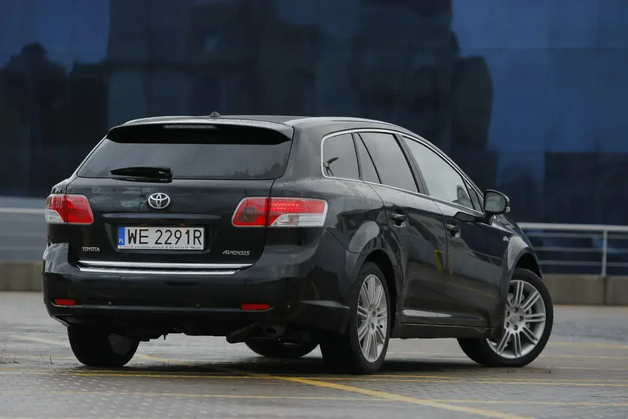 Czarna Toyota Avensis T27, kombi, z widocznym tyłem i tablicą rejestracyjną. Może być pomocna przy wymianie żarówki.