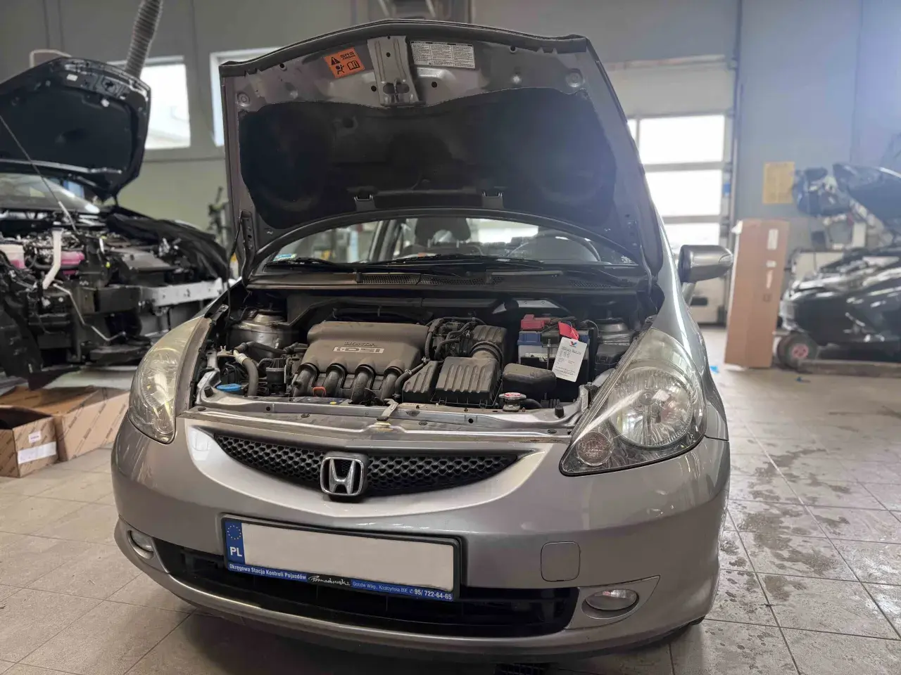 Srebrna Honda Jazz z otwartą maską, widoczny silnik i akumulator. Wymiana żarówki Honda Jazz w warsztacie.