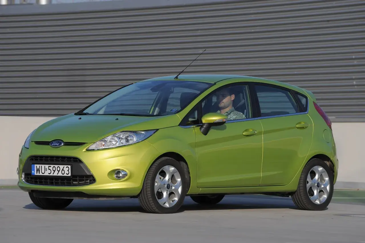 Zielony Ford Fiesta Mk7 z widocznymi żarówkami z przodu, zaparkowany przed budynkiem.