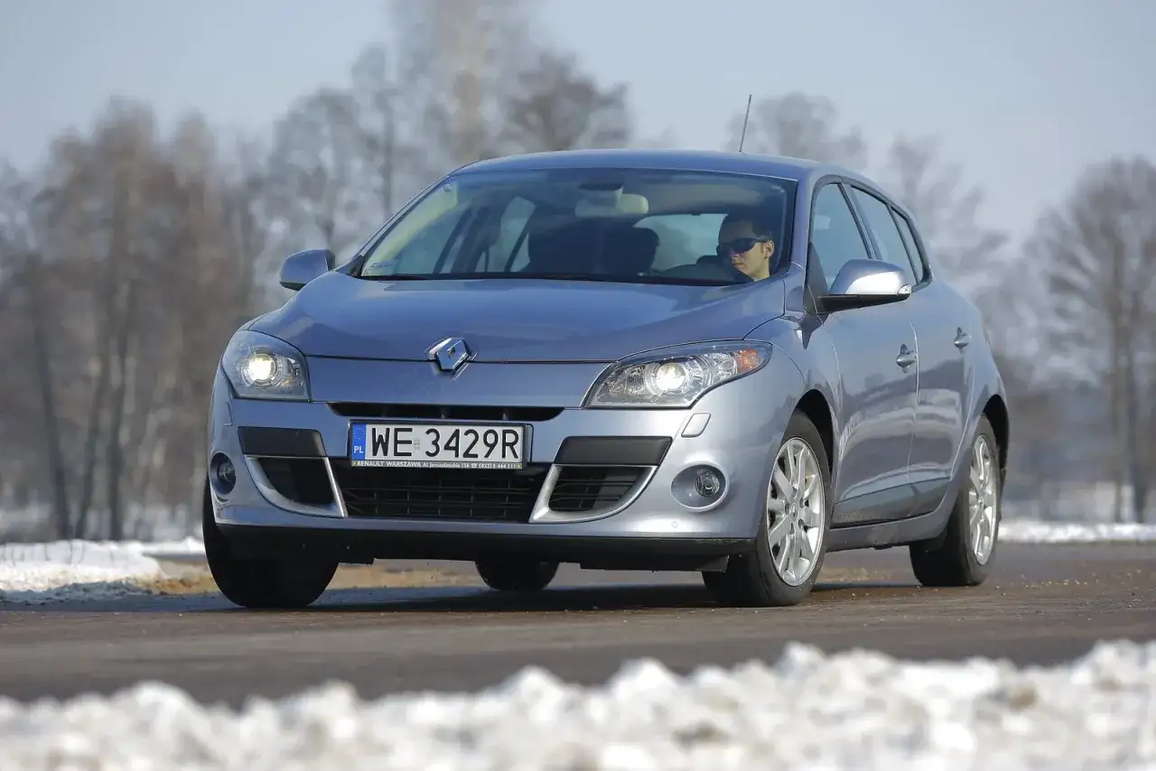 Srebrny Renault Megane 3 z włączonymi żarówkami z przodu, na drodze zimą. Kierowca w okularach.