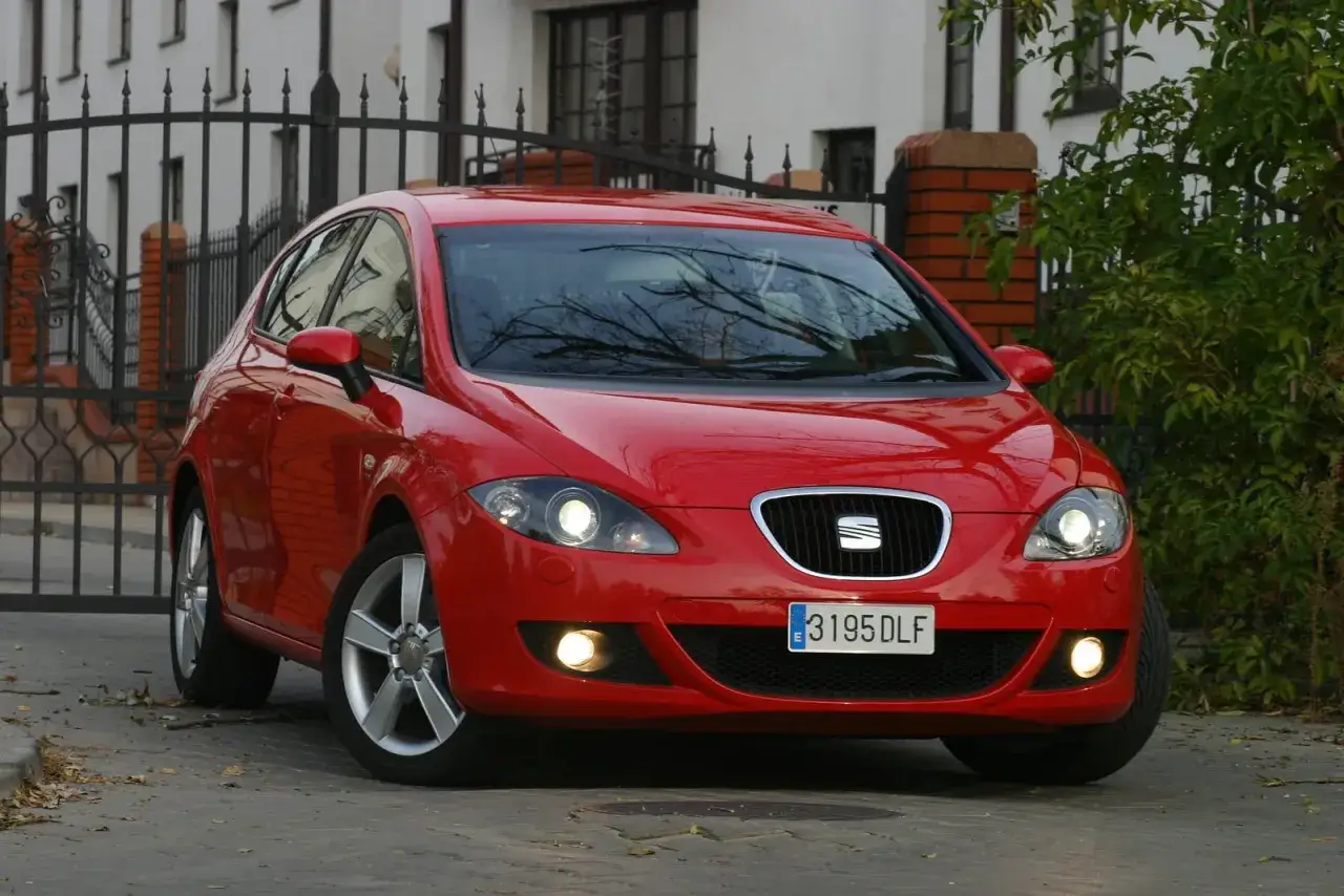 Czerwony Seat Leon 2 z włączonymi światłami, zaparkowany przed bramą.