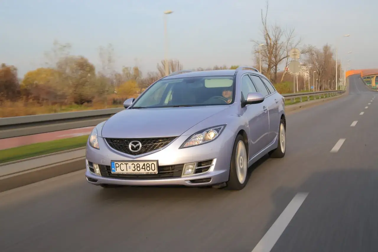 Mazda 6 kombi w kolorze jasnofioletowym jedzie po drodze, jej żarówki mijania oświetlają trasę.