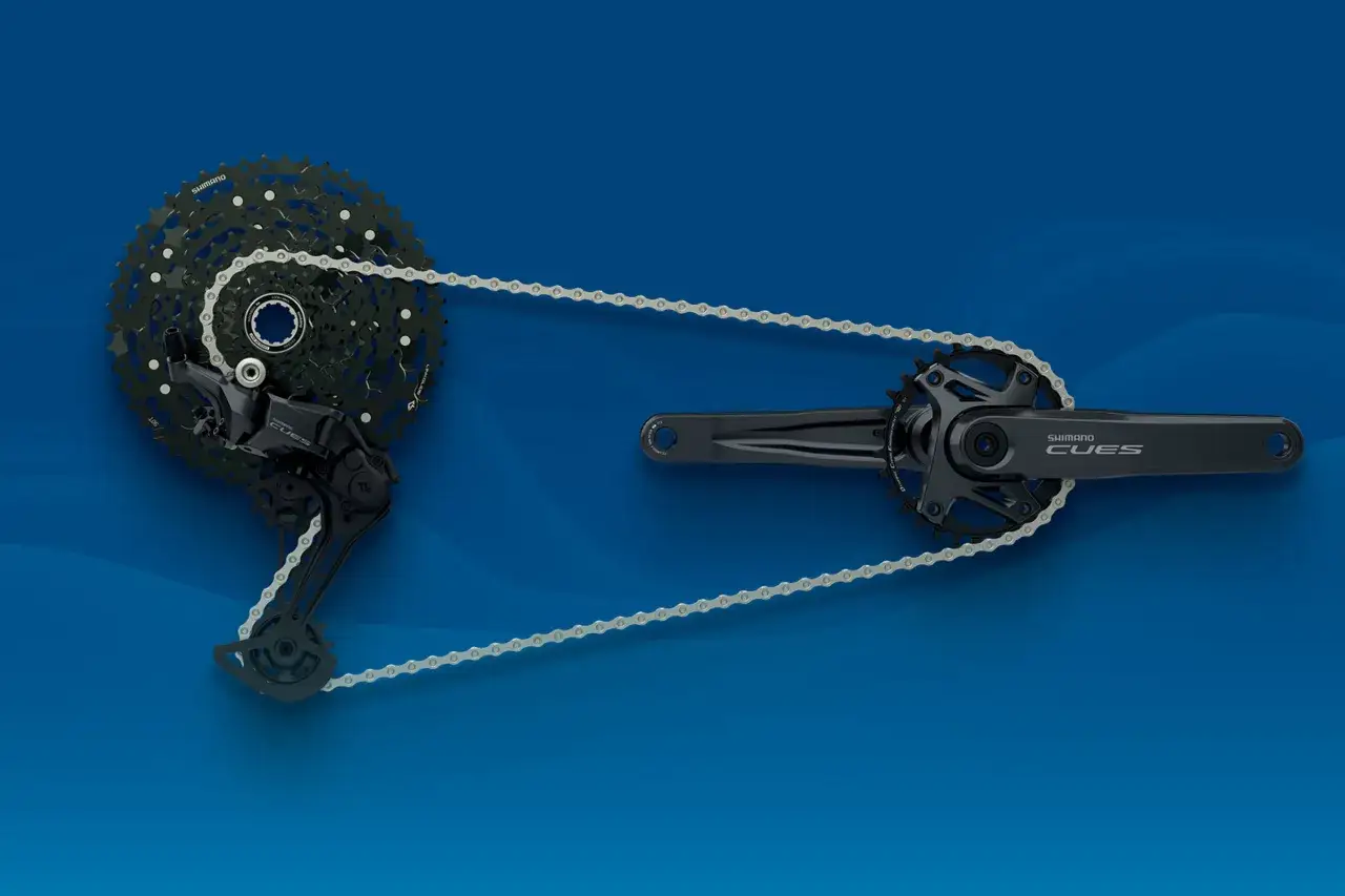Zestaw napędowy Shimano CUES: korba, kaseta i przerzutka tylna połączone łańcuchem, na niebieskim tle.