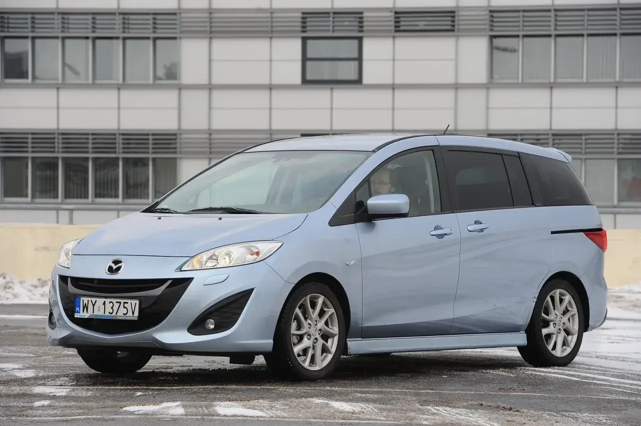 Jasnoniebieska Mazda 5 z widocznymi żarówkami reflektorów, zaparkowana przed nowoczesnym budynkiem.