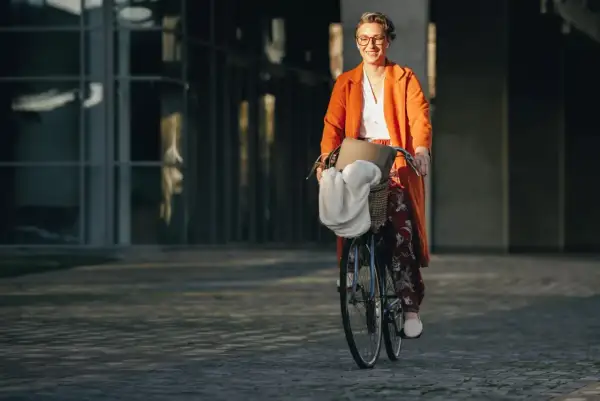 Uśmiechnięta kobieta w pomarańczowym płaszczu jedzie rowerem do pracy. W koszyku ma torbę i biały szal.