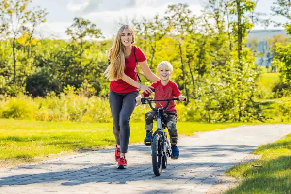 Mama uczy synka, jak nauczyć dziecko jeździć na rowerze. Radosny chłopiec pedałuje, a mama go asekuruje.